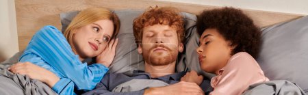 Photo for Love triangle, polyamorous relationship, polygamy, three adults sleeping together, redhead man and multicultural women in pajamas, bedroom, cultural diversity, acceptance, bisexual, banner - Royalty Free Image