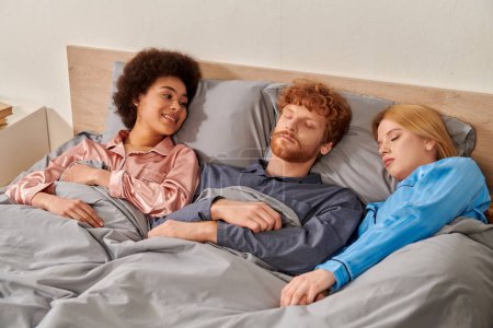 triangle amoureux, heureuse femme afro-américaine regardant somnolent rousse homme près blonde partenaire féminin au lit, polyamour, relations non traditionnelles, multiracial, diversité culturelle 