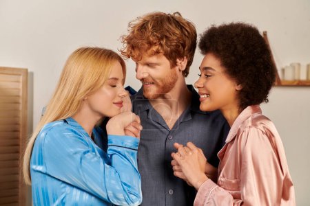 Photo for Polyamory lovers in pajamas, cheerful man with red hair hugging interracial women at home, cultural diversity, non traditional partners, freedom in relationship, acceptance and understanding - Royalty Free Image