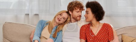 modern family, polygamy concept, freedom in relationship, cultural diversity, redhead man sitting with multicultural women on couch in living room, polyamorous lifestyle, non traditional, banner 
