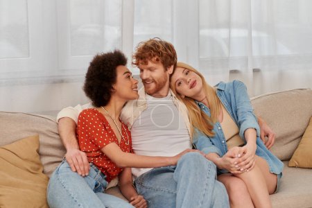 Photo for Polygamy concept, non traditional lovers, freedom in relationship, cultural diversity, redhead man sitting with multicultural women on couch in living room, polyamorous lifestyle, modern family - Royalty Free Image