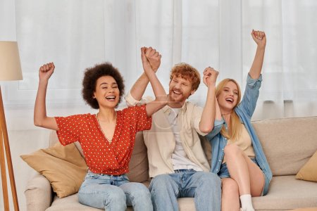 Photo for Polyamorous relationship, cultural diversity, redhead man sitting on couch with happy multiracial female lovers, raised hands, freedom and acceptance, love triangle, people in open relationship - Royalty Free Image