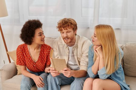 open relationships, redhead man reading book to multicultural women in living room, non traditional lovers, cultural diversity, romance and love, modern family and polygamy concept 