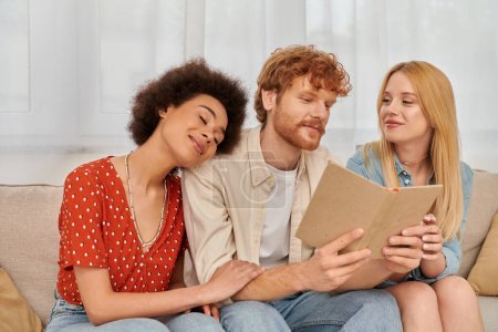 polygamy, open relationships, redhead man reading book to pleased multicultural women in living room, non traditional lovers, cultural diversity, romance and love, modern family concept 
