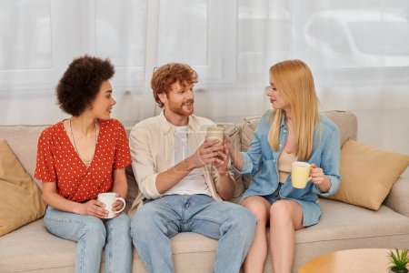 polyamouröse Familie, Beziehungsvielfalt, glückliche Polygamie-Liebhaber, die mit Kaffeebechern auf der Couch sitzen, gemischtrassige Männer und Frauen im Wohnzimmer, bisexuelle und polyamory Menschen 