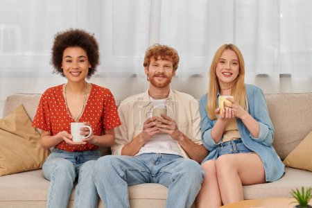 polyamouröses Konzept, Beziehungsvielfalt, glückliche Polygamie-Liebhaber, die auf der Couch sitzen und Kaffeebecher in die Kamera halten, interrassische Männer und Frauen im Wohnzimmer, Bisexualität und Polygamie 