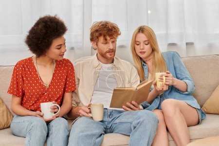 Photo for Alternative relationships, polygamy, intelligent redhead man reading book to multicultural women in living room, modern family, hobby and leisure, polyamorous family, cups of coffee - Royalty Free Image