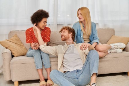 love triangle concept, diversity in relationships and culture, non monogamy, happy redhead man sitting near multicultural women in living room, lovers, modern family, acceptance, open relationship 
