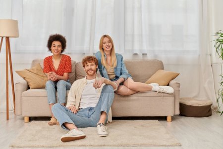 love triangle concept, diversity in relationships and culture, non monogamy, happy redhead man and multicultural women looking at camera in living room, lovers, acceptance, open relationship 