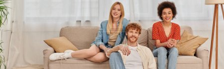Photo for Love triangle concept, diversity in relationships and culture, non monogamy, happy redhead man and multicultural women looking at camera in living room, lovers, acceptance, open relationship, banner - Royalty Free Image