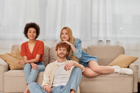 Photo for Polygamy concept, diversity in relationships and culture, non monogamy, happy redhead man and multicultural women looking at camera in living room, lovers, acceptance, open relationship, banner - Royalty Free Image