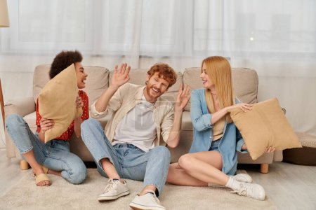Photo for Polygamy, acceptance and bonding, group relationship, happy multicultural women and bearded man fighting with pillows, open relationship, diversity and bonding, non monogamy, three people - Royalty Free Image