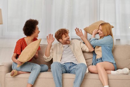 polyamorous family, group relationship, cheerful multicultural women and bearded man fighting with pillows, open relationship, diversity and bonding, non monogamy, three people 