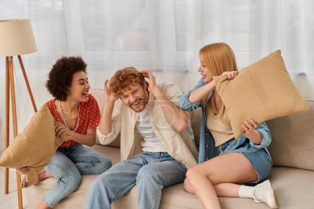 Photo for Polyamorous family, love triangle, cheerful multicultural women and bearded man fighting with pillows, open relationship, diversity and bonding, non monogamy, three people - Royalty Free Image