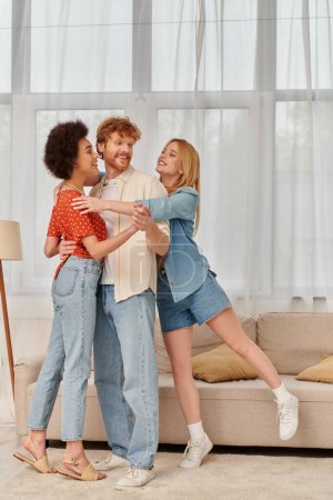 alternative family, happy multicultural women hugging redhead man in living room, cultural diversity, freedom in relationships, love triangle, bisexual and free spirited people 