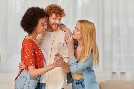 alternative family, happy multicultural women near redhead man in living room, interracial lovers, cultural diversity, freedom in relationships, love triangle, bisexual and free spirited people 