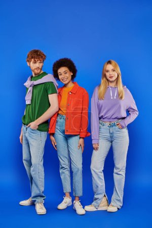 Photo for Polygamy concept, happy multicultural women and redhead man looking at camera on blue background, studio photography, cultural diversity, polygamy, modern family, colorful clothes - Royalty Free Image