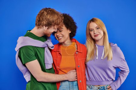 polyamory concept, cheerful redhead man hugging african american woman near blonde lover on blue background, studio photography, cultural diversity, polygamy, modern family, understanding 