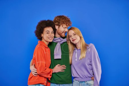 Photo for Polyamory concept, cheerful multicultural women looking at camera and hugging with redhead man on blue background, studio photography, cultural diversity, polygamy, young lovers in casual attire - Royalty Free Image