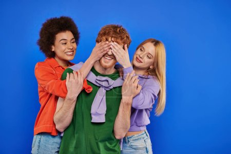 Photo for Polyamory, three people, beautiful multiracial women covering eyes of young redhead man on blue background, studio shot, vibrant colors, casual attire, modern family, alternative relationships, - Royalty Free Image