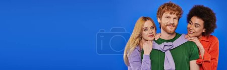 alternative relationships, polygamy, portrait of three people, young redhead man and beautiful multiracial women on blue background, studio shot, vibrant clothes, polyamory, modern family, banner 