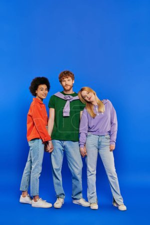 Photo for Alternative family, polygamy, portrait of three people, young redhead man and beautiful multiracial women on blue background, studio shot, vibrant clothes, polyamory, modern relationships - Royalty Free Image