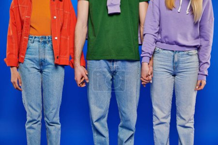 alternative family, cropped view of three polygamy people, young man and women holding hands on blue background, studio shot, vibrant clothes, polyamory, modern relationships 