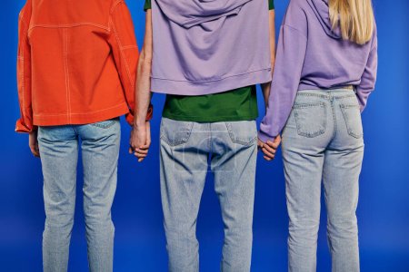 Photo for Polygamy, back view of polyamory three people, young man and women holding hands on blue background, studio shot, vibrant clothes, love triangle, cropped shot, bonding, alternative relationships - Royalty Free Image