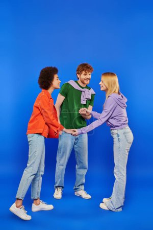 Photo for Polyamorous family concept, polygamy lovers, young man and bisexual multicultural women holding hands on blue background, studio shot, denim fashion, love triangle, bonding and acceptance - Royalty Free Image