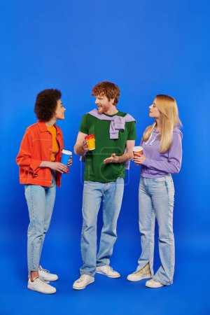 polygamy people with coffee, young redhead man and beautiful multiracial women holding paper cups on blue background, studio shot, takeaway drink, polyamorous lovers, conversation 