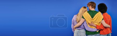 Photo for Love triangle, polyamory, three people, back view of redhead man hugging multiracial women on blue background, studio shot, vibrant colors, casual clothes, stylish attire, modern family, banner - Royalty Free Image