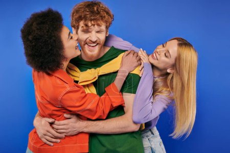 alternative relationships, polyamory lovers, cheerful multiethnic women kissing and hugging young redhead man on blue background, studio shot, vibrant colors, modern family, bonding and acceptance 
