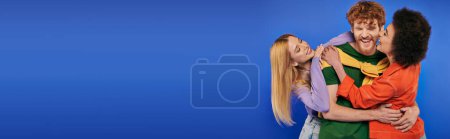 alternative relationships, polyamory lovers, cheerful multiethnic women kissing and hugging young redhead man on blue background, studio shot, vibrant colors, modern family, acceptance, banner