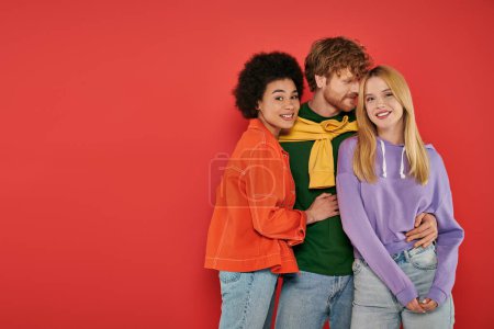 polyamorous relationship concept, polygamy lovers, young man and multicultural women hugging on coral background, studio shot, denim fashion, love triangle, bonding and acceptance 