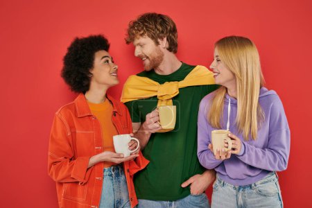 polyamouröses Konzept, junge multikulturelle Frauen und rothaarige Männer mit Tassen auf Korallengrund, Studioaufnahme, lebendige Farben, Morgenroutine, Kaffee, glückliche Polygamie-Liebhaber 