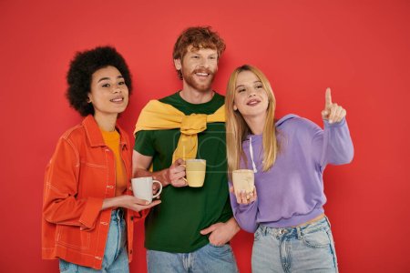 open relationships concept, young blonde woman pointing away near redhead man, holding cups on coral background, vibrant colors, morning routine, coffee, happy polygamy lovers, polyamorous  