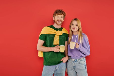 cheerful young couple holding cups of coffee on coral background, morning routine, vibrant colors, stylish outfits, modern family, people holding mugs with hot drink, husband and wife 