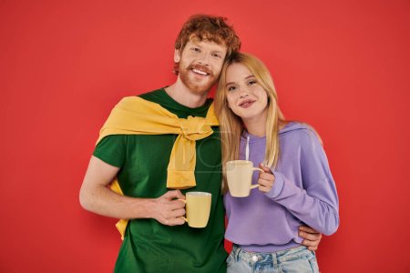 positive young couple holding cups of coffee on coral background, morning routine, vibrant colors, stylish outfits, modern family, people holding mugs with hot drink, husband and wife 