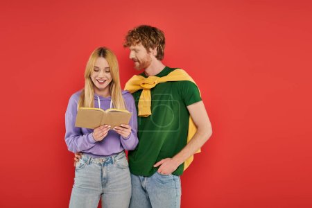 young man looking at girlfriend reading book on coral background, vibrant colors, stylish outfit, youth and intelligence, blonde woman and redhead man spending lovely time together, romance 