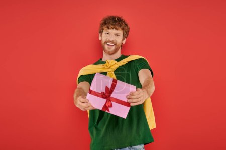 Urlaub, fröhlicher rothaariger Mann mit Bart posiert in lässiger Kleidung auf Korallengrund, hält Geschenkschachtel, festliche Anlässe, verpacktes Geschenk, Mode und Trend, urbaner Stil, Glück 
