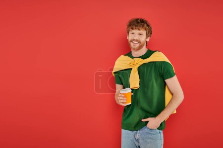 morning coffee, redhead man with beard and curly hair holding paper cup on coral background, vibrant colors, male fashion, takeaway drink, happy and stylish man posing with hand in pocket 