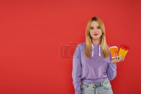 takeaway drink, youthful fashion, blonde young woman in casual attire holding paper cups on coral background, cheerful, looking at camera, vibrant colors, fashion forward, hot beverage 