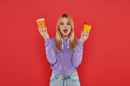 Photo for Takeaway drink, youthful fashion, blonde young woman in casual attire holding paper cups on coral background, choice, open mouth, looking at camera, vibrant colors, fashion forward, hot beverage - Royalty Free Image
