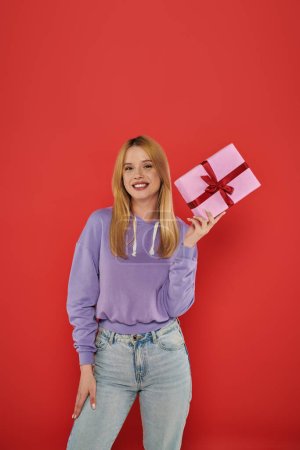 holiday, positivity, blonde young woman in casual attire holding present on coral background, looking at camera, vibrant colors, wrapped gift box, attractive and stylish, festive occasions 