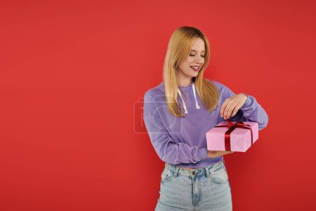 holiday, happiness, blonde young woman in casual attire opening present on coral background, vibrant colors, wrapped gift box, attractive and stylish, festive occasions, purple hoodie and jeans 