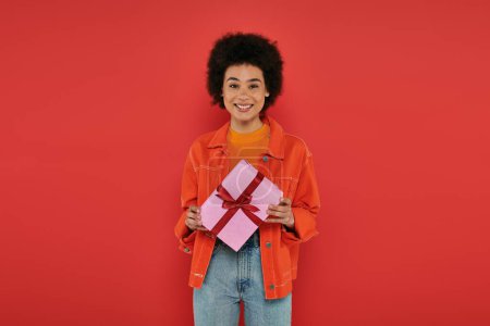 Urlaub, glücklich afrikanisch-amerikanische Frau in lässiger Kleidung hält eingewickelt Geschenk auf Korallenhintergrund, lebendige Farben, Geschenkbox, attraktive und stilvolle, festliche Anlässe, Blick in die Kamera 