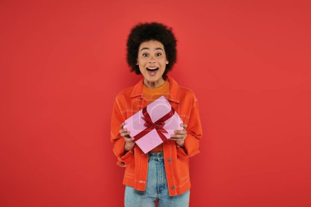 Urlaub, erstaunt afrikanisch-amerikanische Frau in lässiger Kleidung halten verpackt Geschenk auf Korallenhintergrund, lebendige Farben, Geschenkbox, attraktive und stilvolle, festliche Anlässe, Blick in die Kamera 