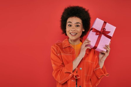 Urlaub, positive afrikanisch-amerikanische Frau in lässiger Kleidung mit eingewickelten Geschenk auf Korallenhintergrund, lebendige Farben, Geschenkbox, attraktive und stilvolle, festliche Anlässe, Blick in die Kamera