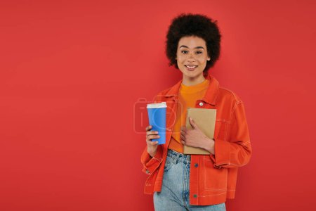 boisson à emporter, femme afro-américaine gaie en tenue décontractée tenant livre sur fond de corail, couleurs vives, attrayant et élégant, café à emporter, tasse en papier, étudiante regardant la caméra 