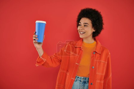 takeaway drink, joyful african american woman in casual attire looking at paper cup on coral background, vibrant colors, attractive and stylish, coffee to go, disposable cup with hot beverage 